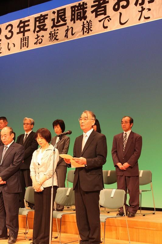 若泰参事がマイクの前で歌を熱唱し、その横で他の退職者達が歌を聞いている写真